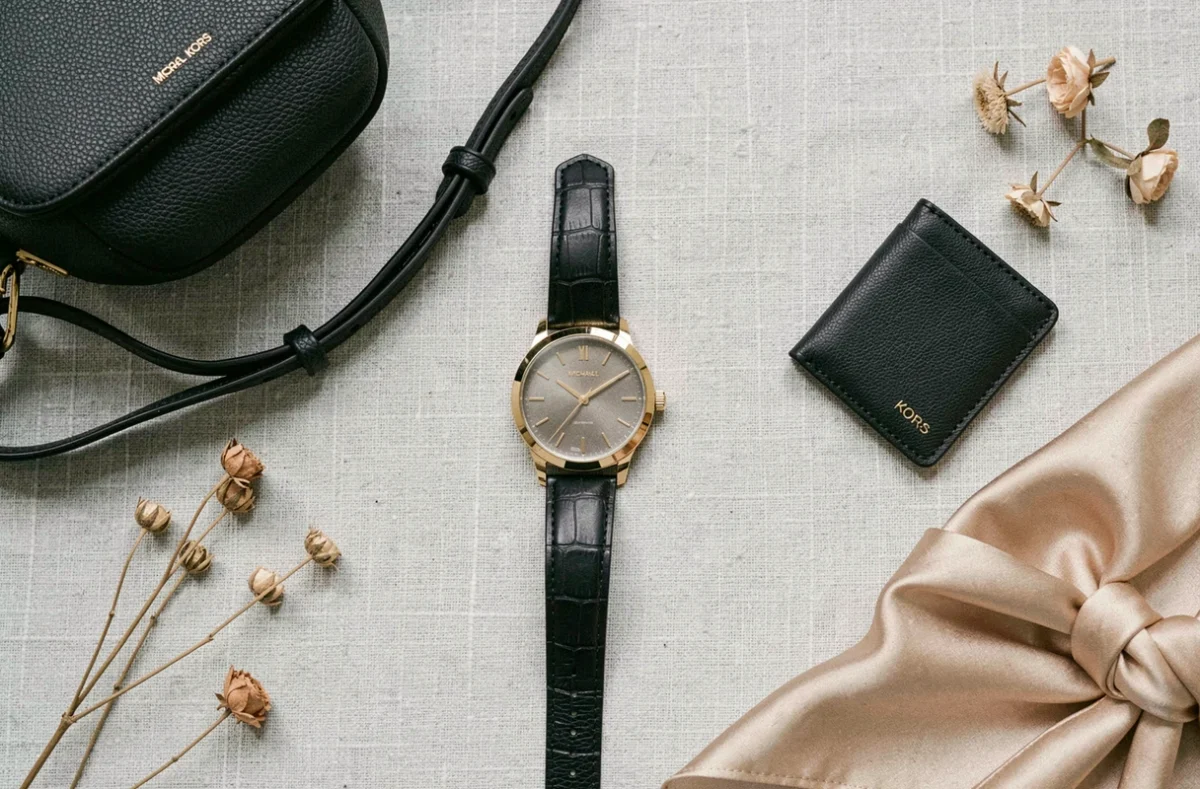 Flat lay de produits Michael Kors incluant une montre dorée, un sac bandoulière noir et un porte-cartes sur fond lin gris